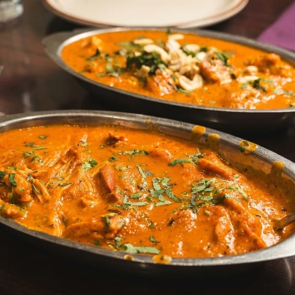 Two oval metal dishes filled with orange curry, garnished with cilantro and cashews, on a wooden table.