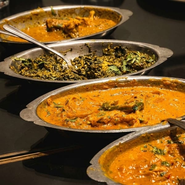 Four oval metal dishes filled with various Indian curries, including green and orange sauces.